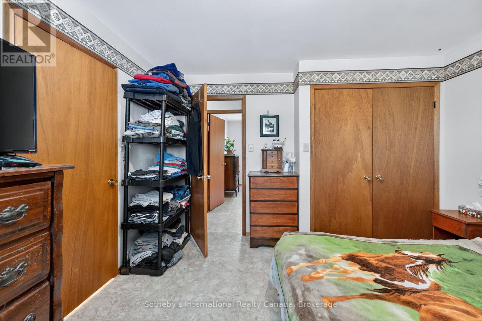 Bedroom with wooden closet doors, dressers, and an open rack of folded clothes beside a horse-print bedspread.