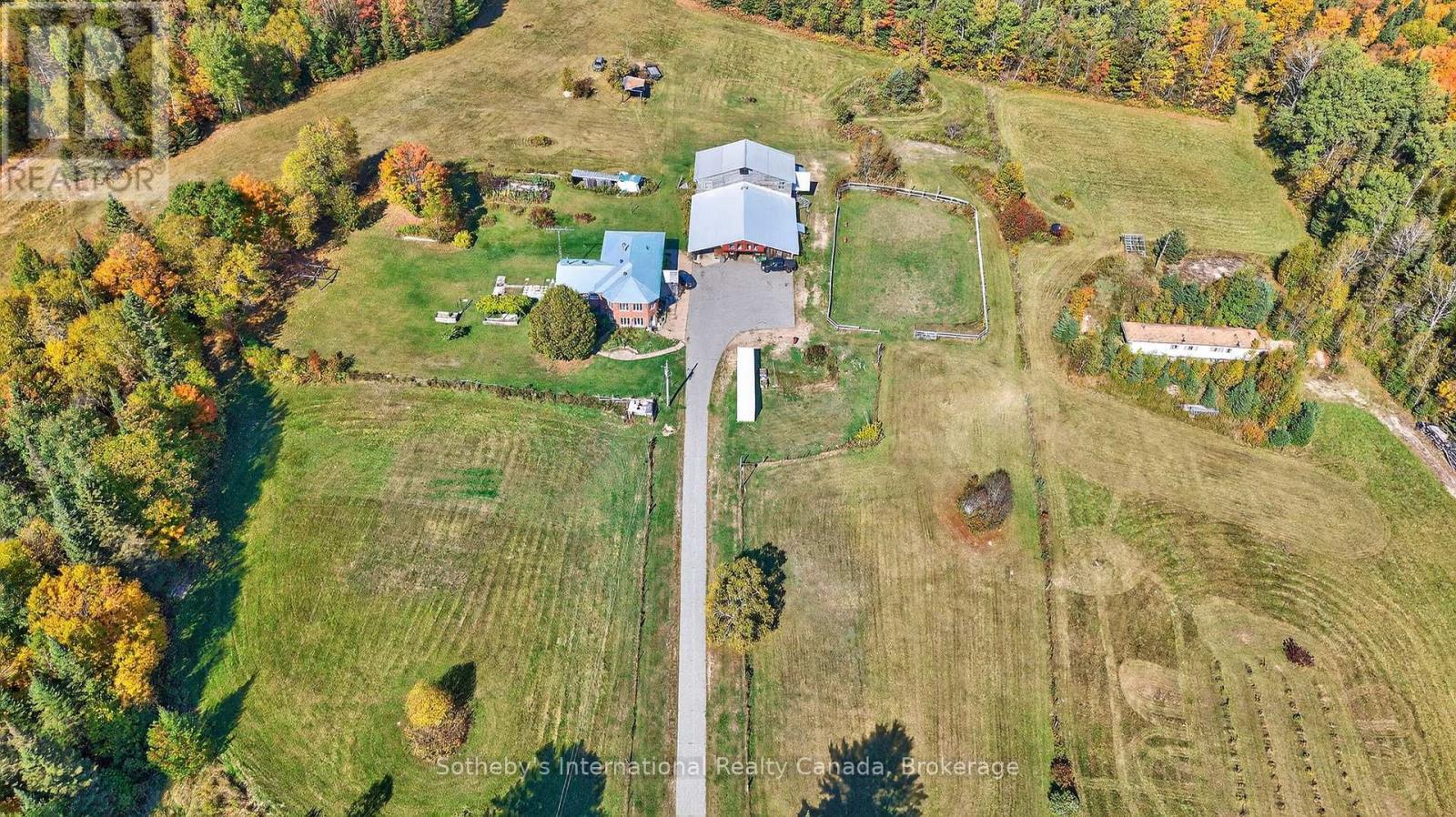 Aerial view of a rural property with multiple outbuildings, open fields, fenced paddocks, and autumn woodland.