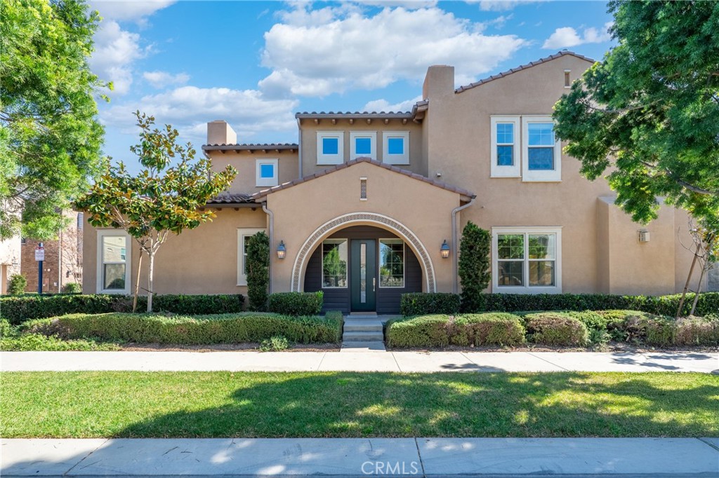 Two-story tan stucco home with an arched entryway, dark double doors, and landscaped front lawn.