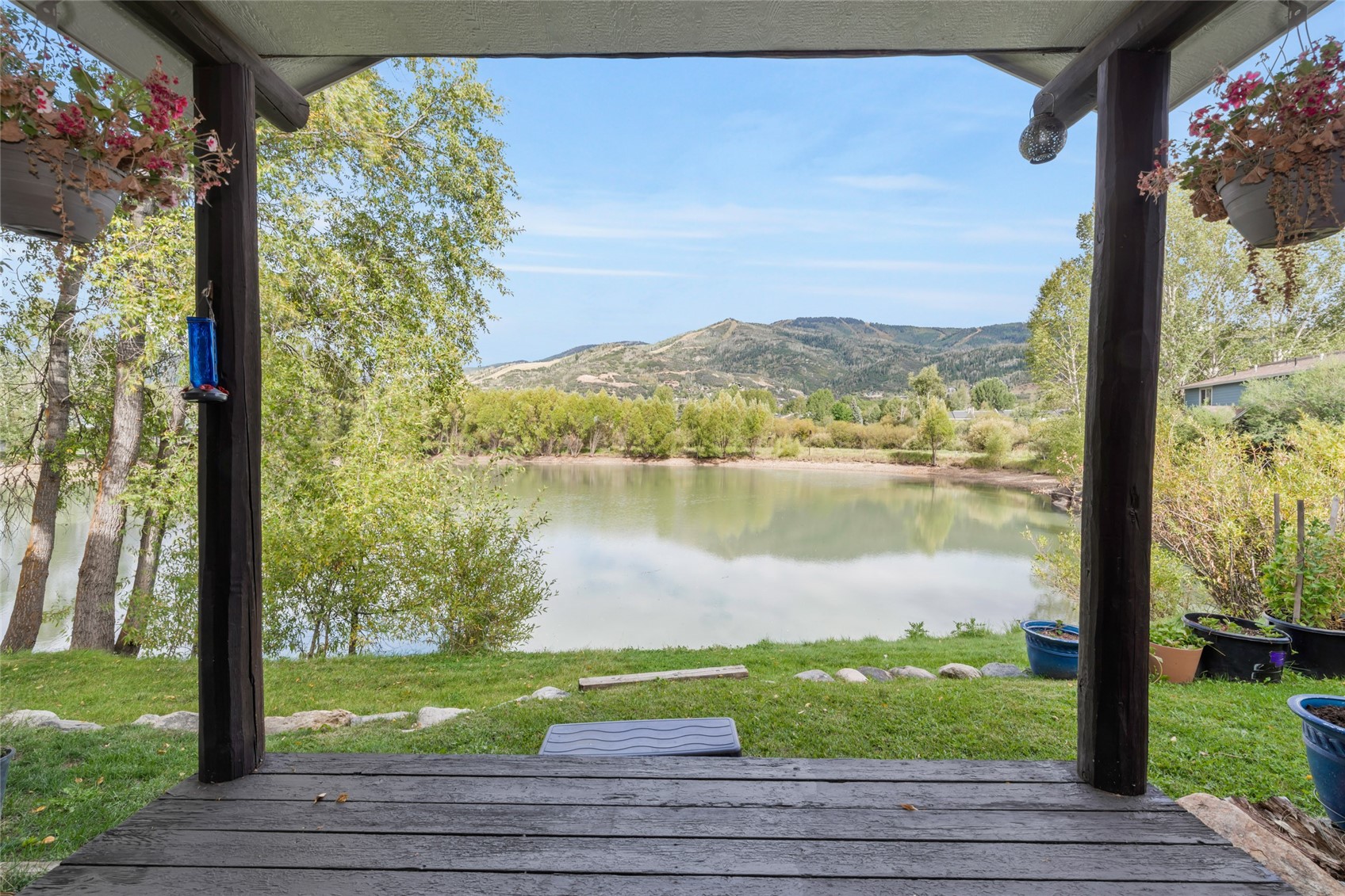 Covered deck overlooking a pond and tree-covered mountains, framed by hanging flowers and potted plants.