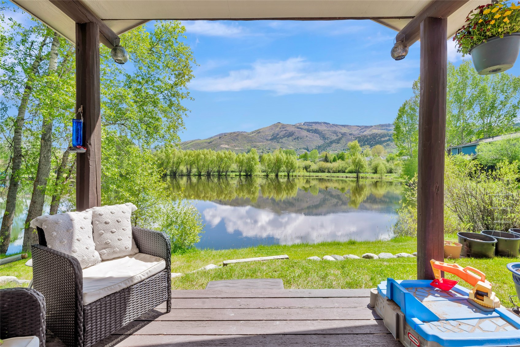 Covered deck with wicker seating overlooking a reflective pond and mountain ridge.