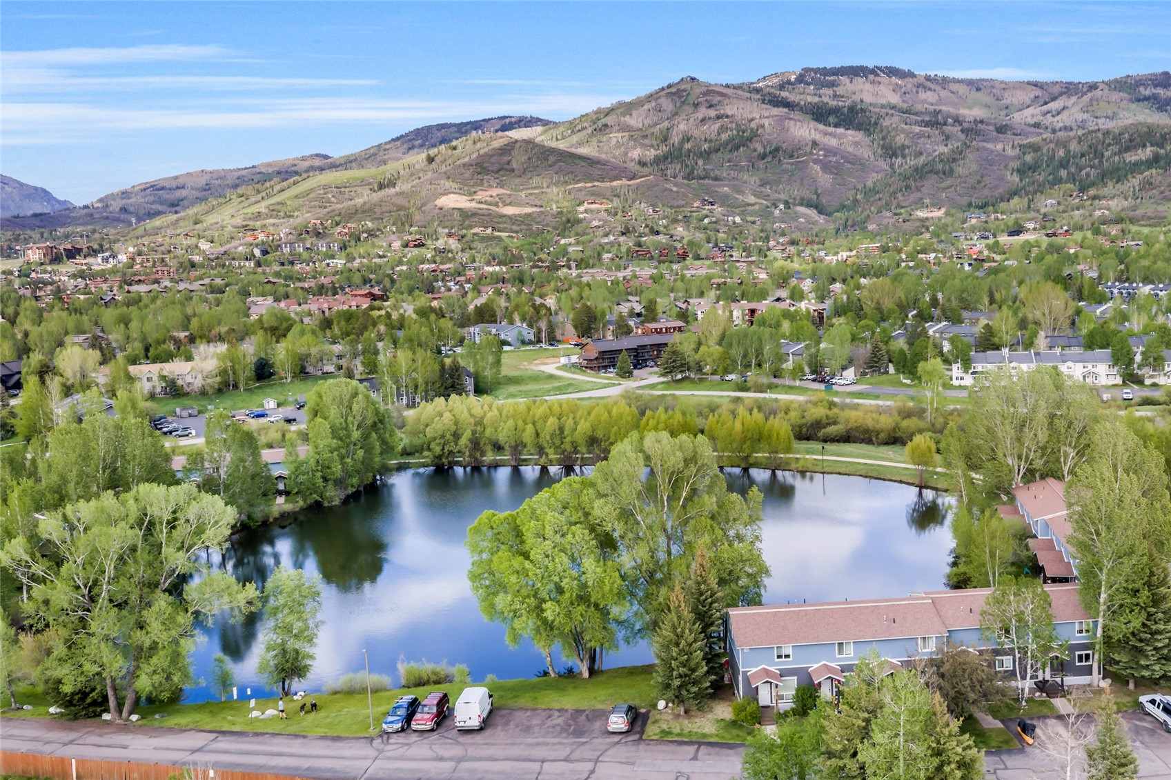 Aerial view of a pond bordered by homes, with a mountain town and hills beyond.