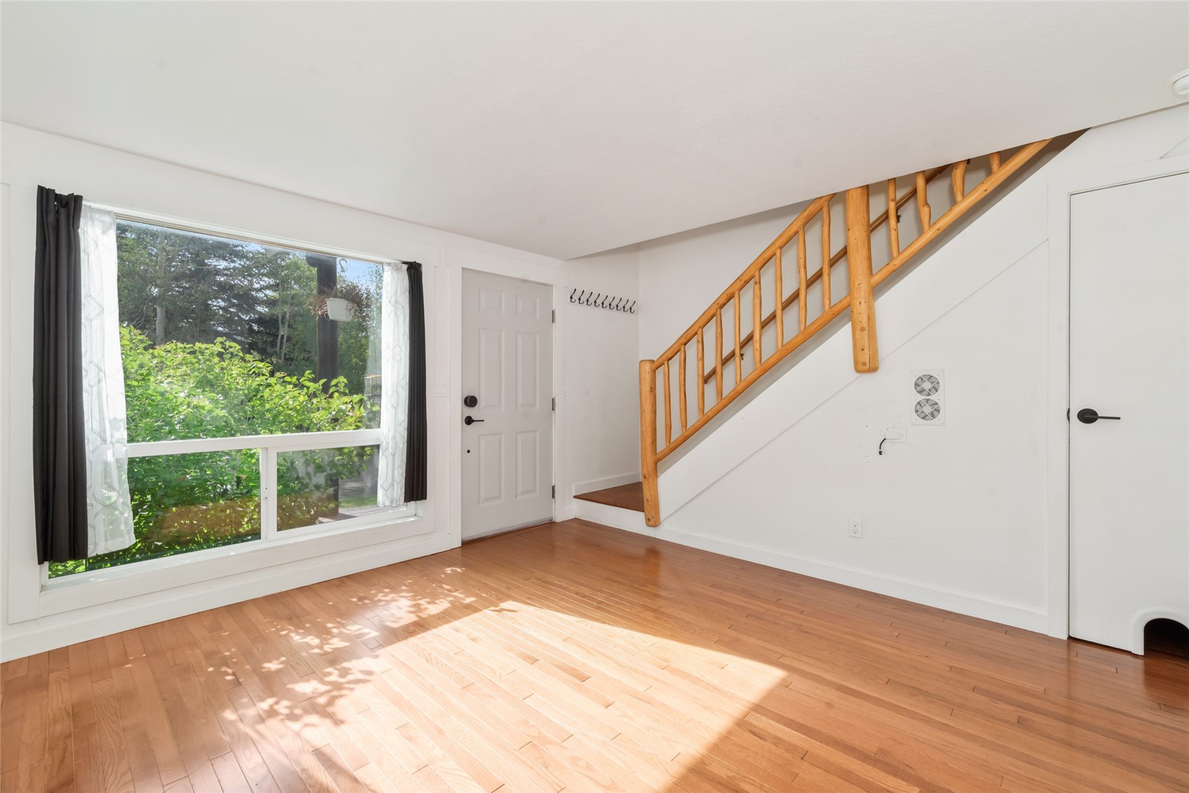 Vacant living room with hardwood floors, a large window, and a wooden staircase.