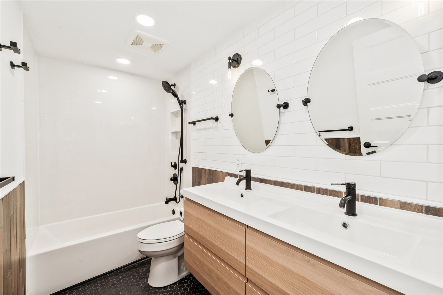 Bathroom with a white tub and shower beside a dual-sink wood vanity and two oval mirrors.