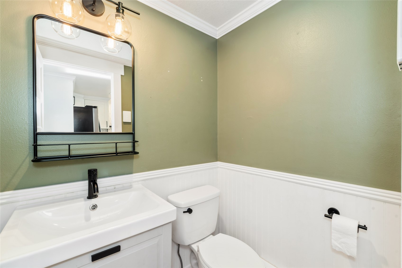 Bathroom with a white vanity and toilet, green walls, white beadboard, and a black-framed mirror.