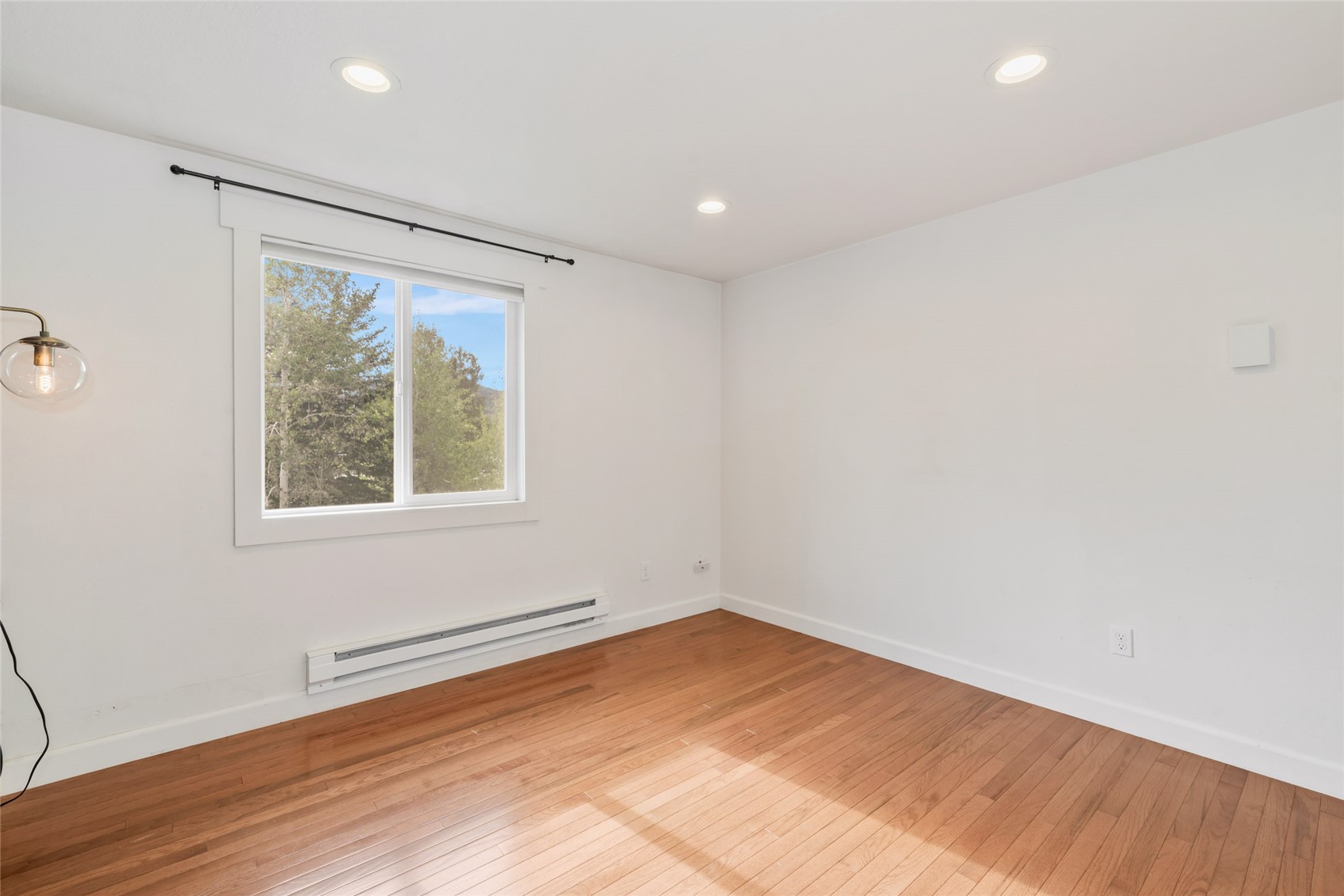 Empty room with hardwood floors, recessed lights, and a window overlooking trees.