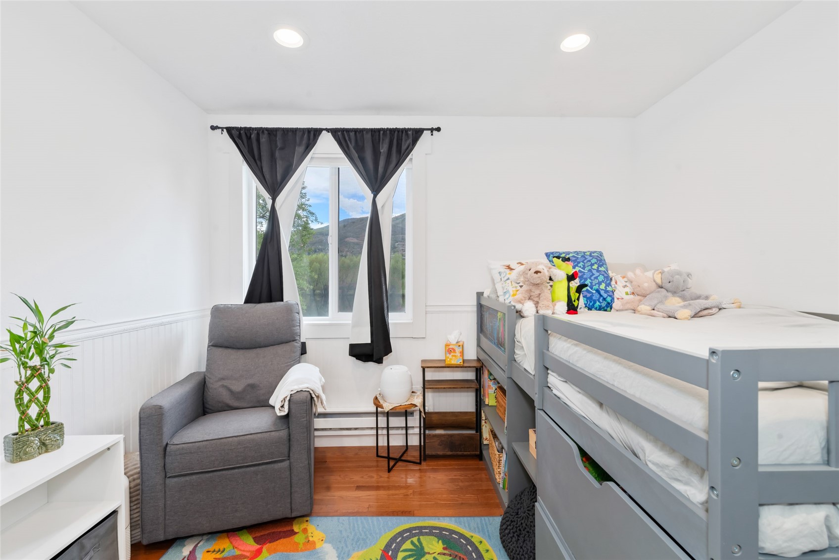 Bedroom with a gray bunk bed, stuffed animals, and a recliner beside a mountain-view window.