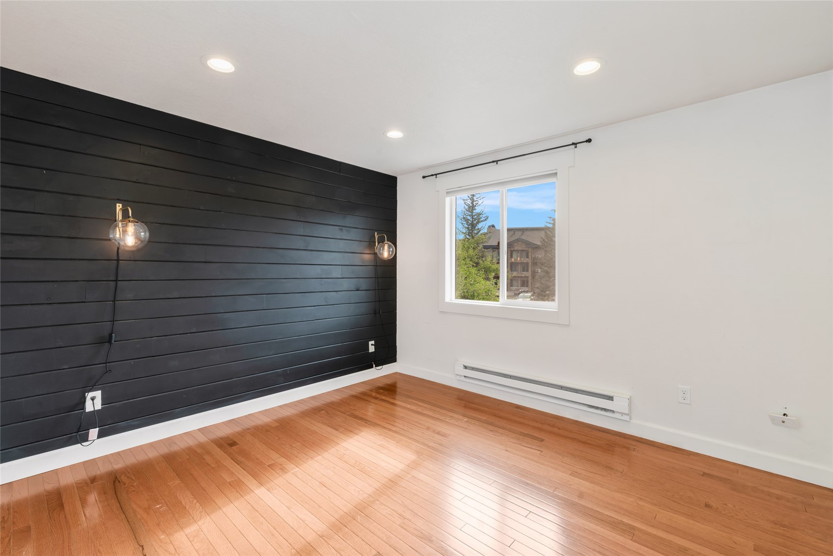 Empty room with hardwood floors, a black accent wall, and a window overlooking a neighboring building.
