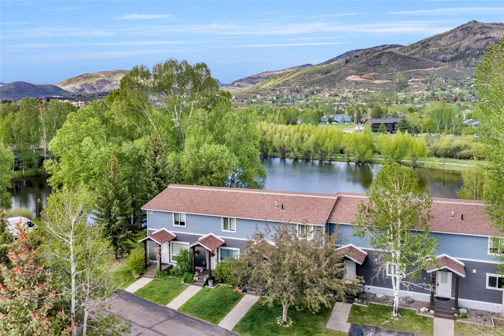 Blue townhomes beside a pond, with leafy trees and mountains behind them.