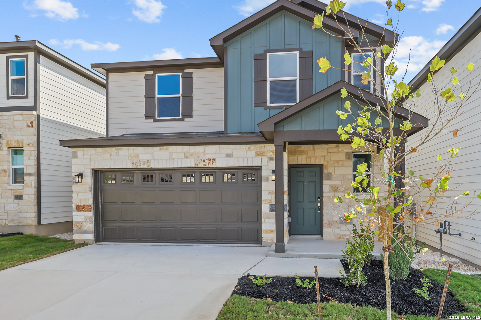 Two-story home with stone and blue-green siding, a double garage, and a young tree in the front yard.