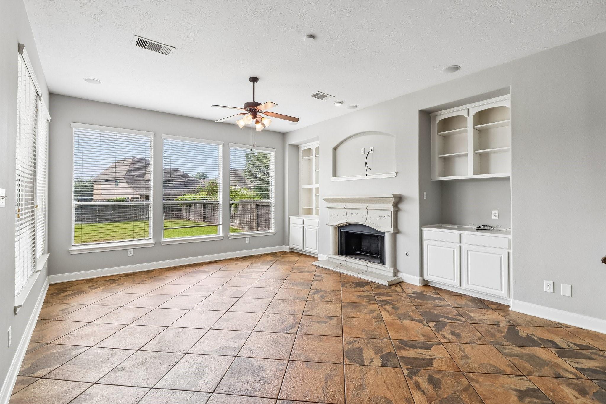 Empty living room with a decorative fireplace, built-in cabinets, large windows, and patterned tile flooring.