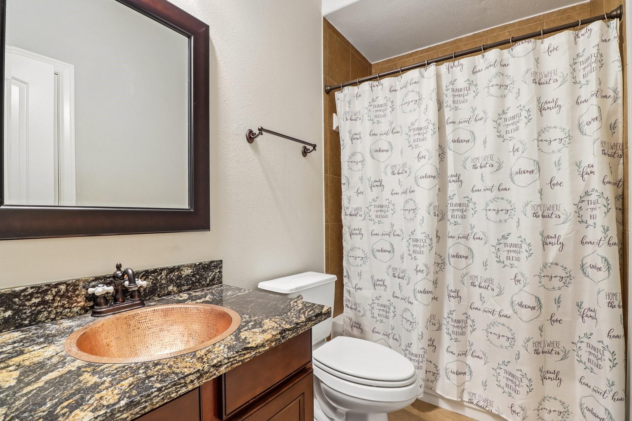Bathroom with copper-toned vessel sink, granite vanity, and printed white shower curtain.