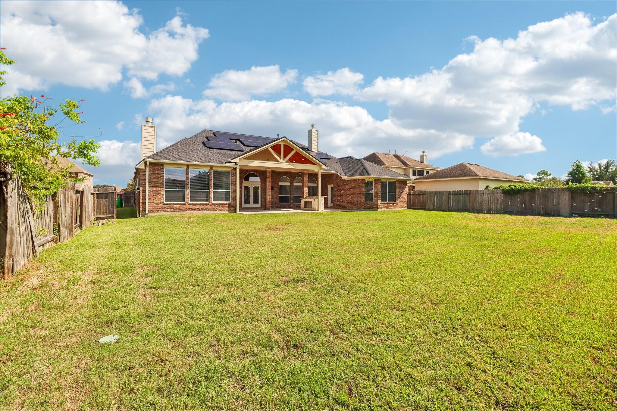 Brick home’s rear exterior with solar panels, covered patio, and a large fenced lawn.