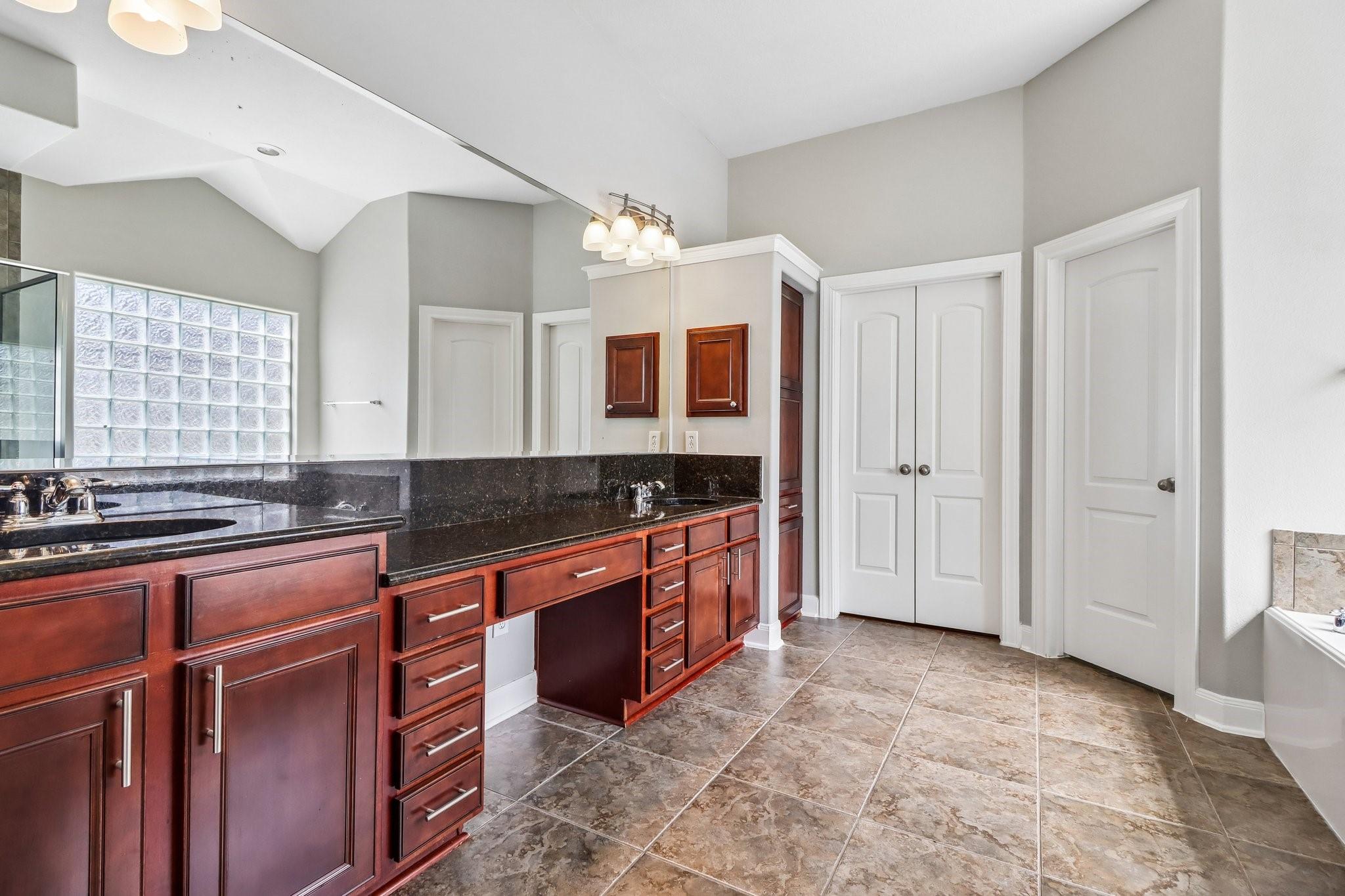 Bathroom vanity with dark granite counters, reddish wood cabinets, and a glass-block window.