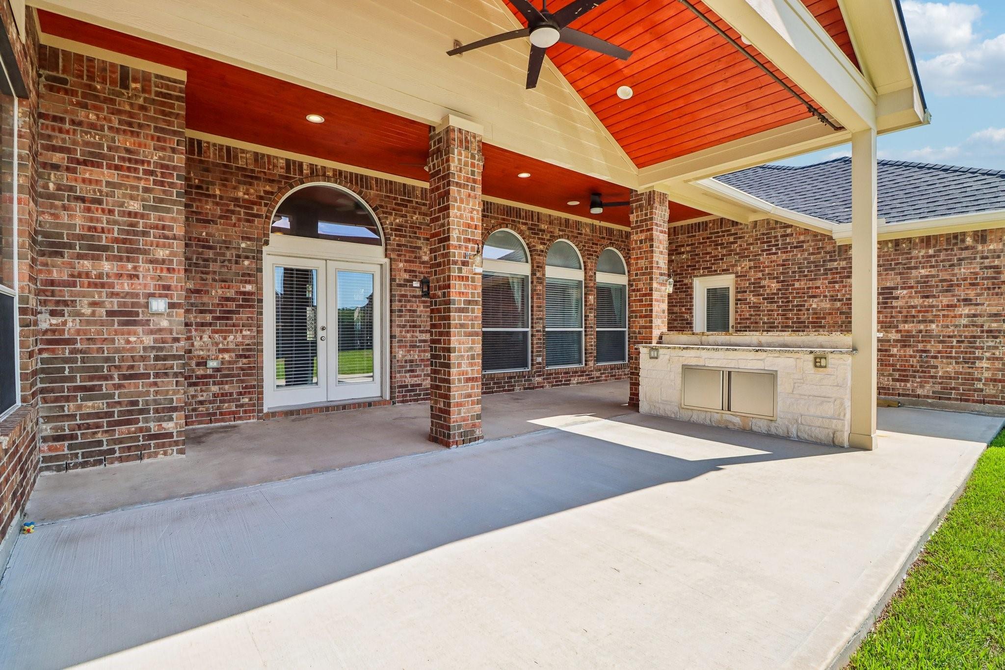 Covered brick patio with a wood-paneled ceiling, ceiling fan, and stone outdoor kitchen island.