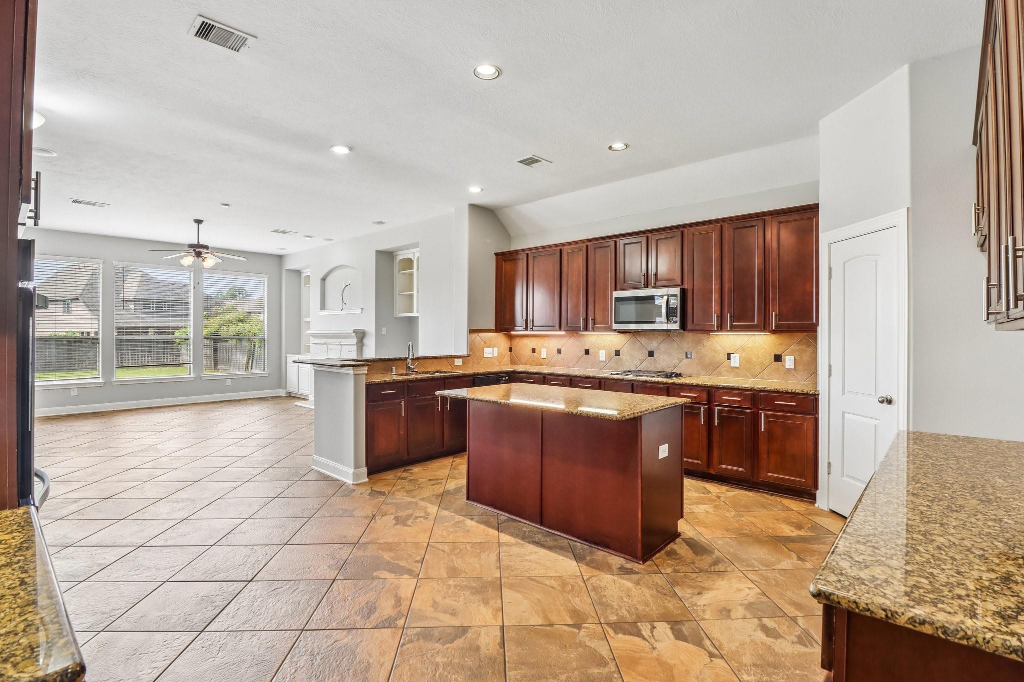Vacant kitchen with dark wood cabinets, granite island, tan tile floors, and large windows.
