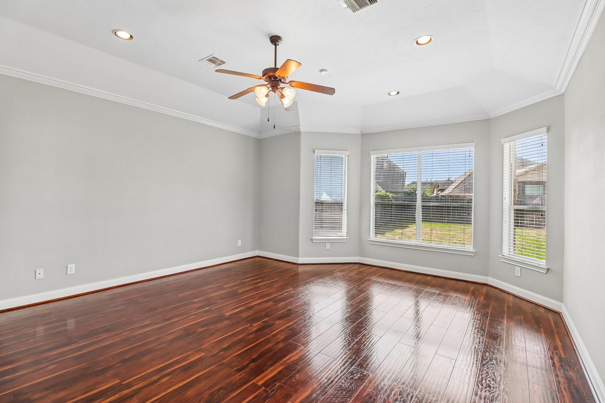 Empty room with glossy dark wood floors, three windows, and a ceiling fan with lights.