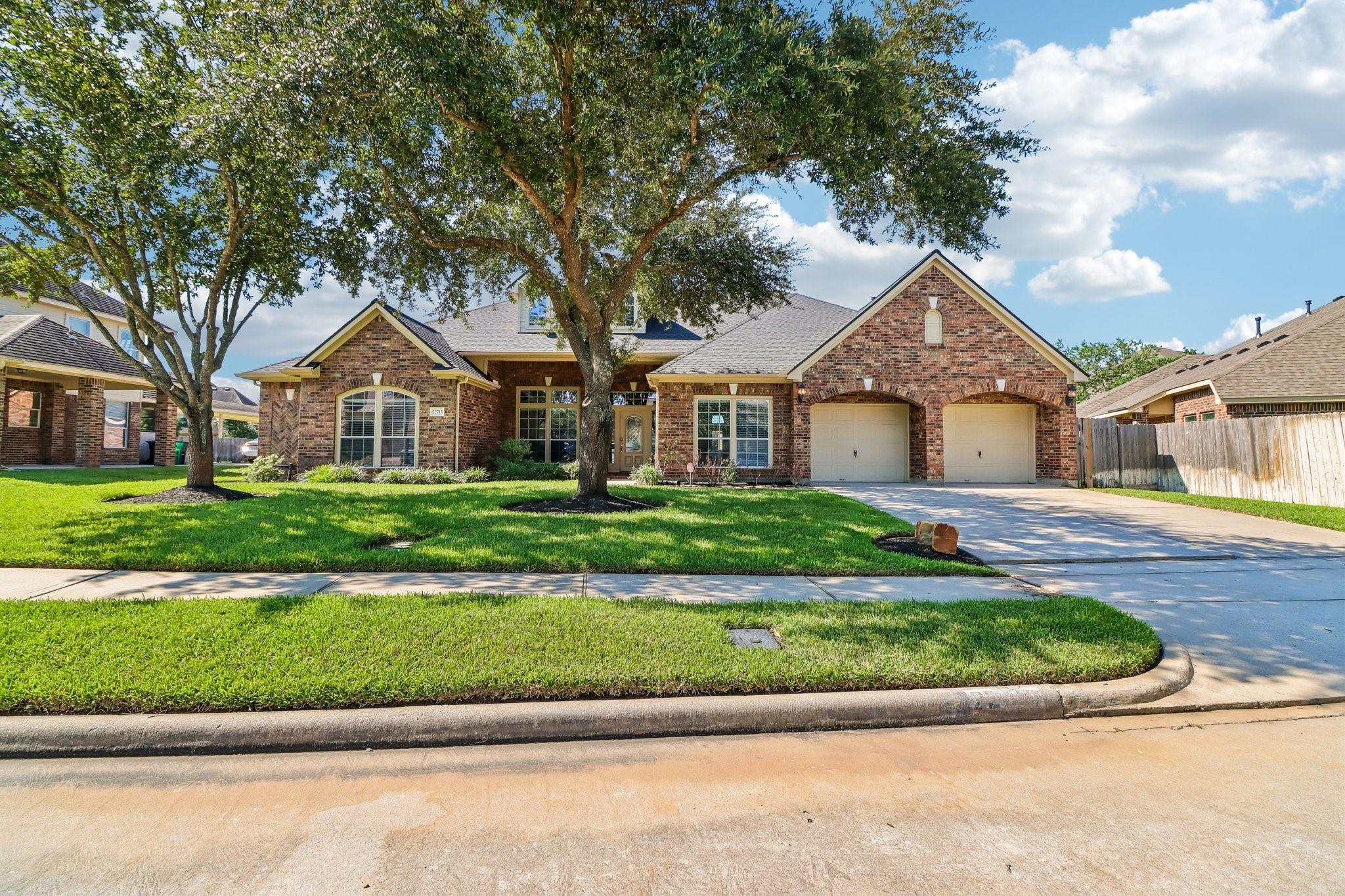 One-story brick house with a two-car garage, mature shade trees, and a broad green lawn.