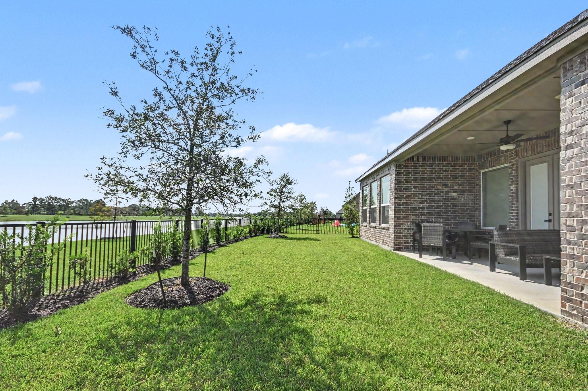 Brick home’s covered patio and lawn beside a fenced pond, with outdoor seating and young trees.