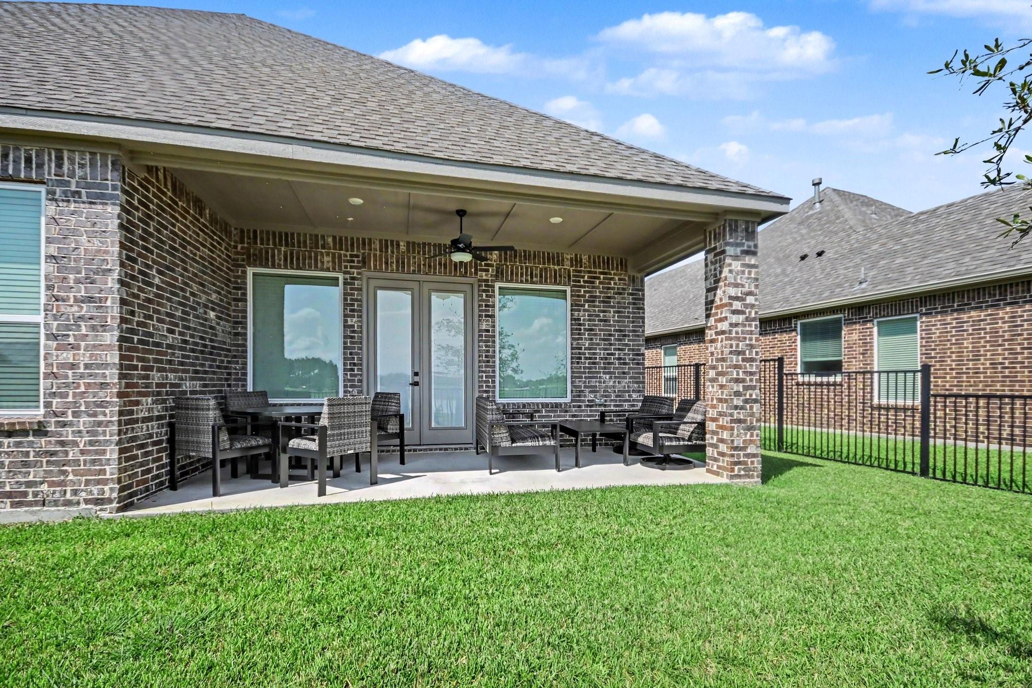 Covered brick patio with outdoor seating and ceiling fan overlooking a green lawn.