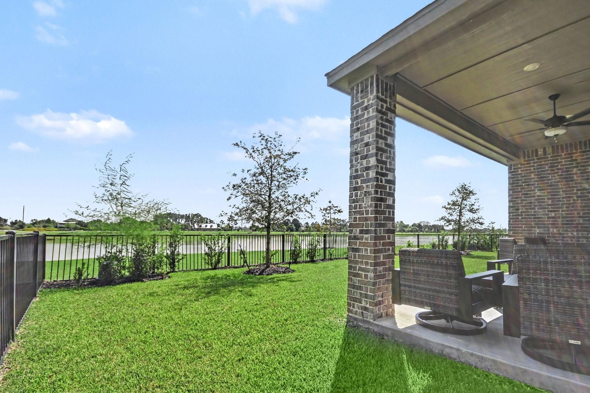 Covered brick patio with wicker seating overlooking a fenced lawn and pond.