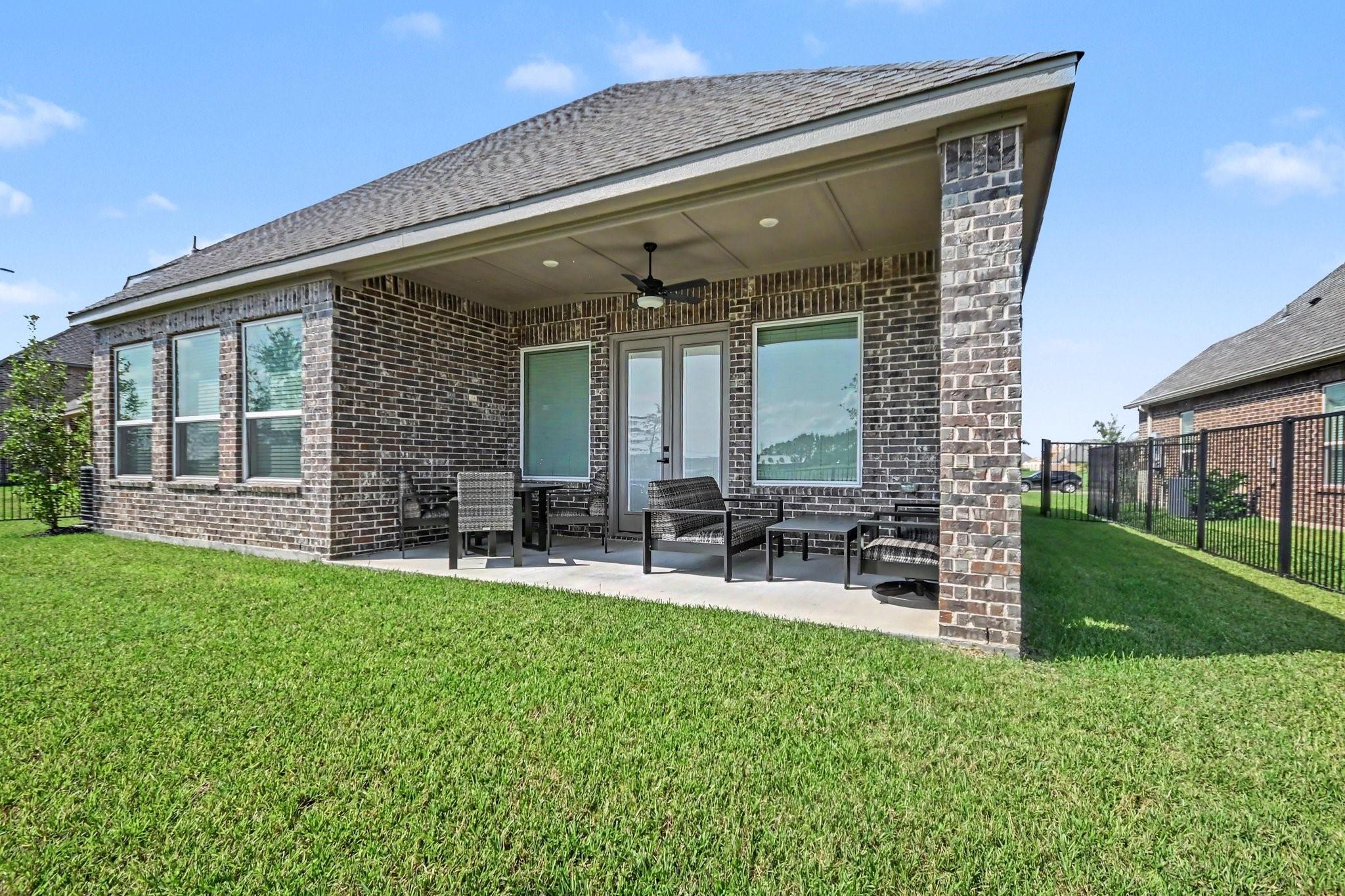 Brick home’s covered rear patio with outdoor seating, ceiling fan, and a broad green lawn.