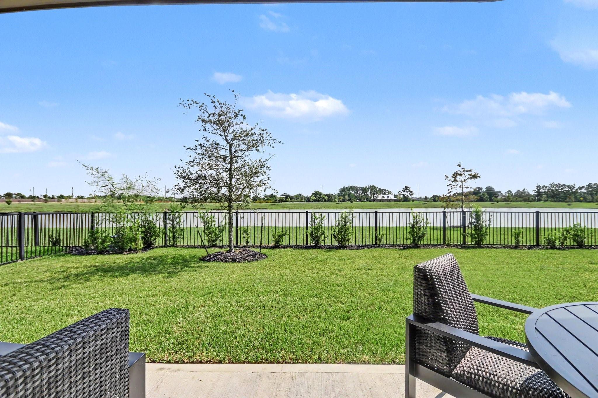 Backyard patio overlooking calm water, with a broad lawn, black fence, and wicker seating.