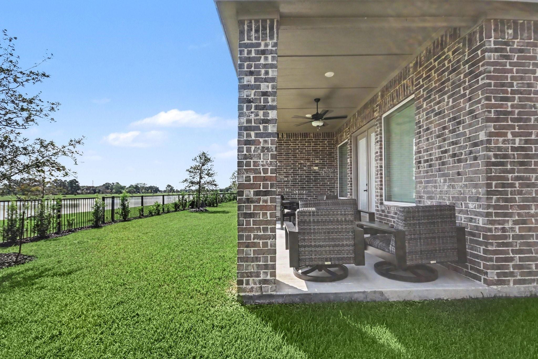 Covered brick patio with wicker seating overlooks a fenced lawn and water.