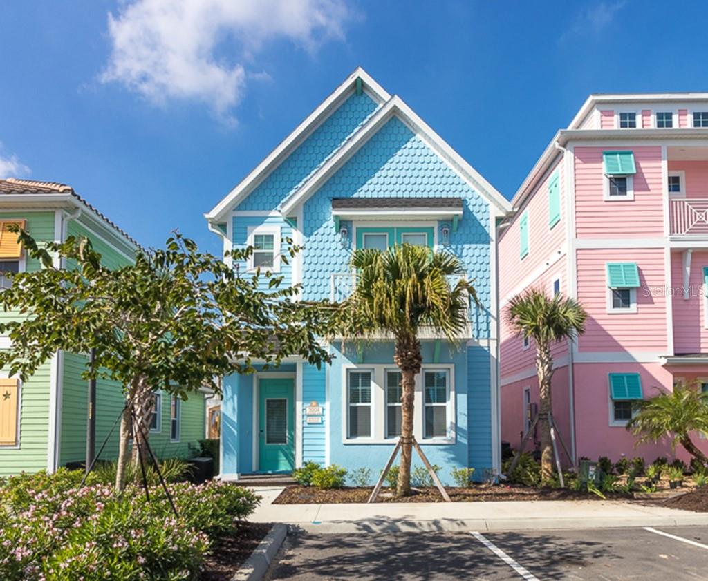 Blue two-story home with a steep gabled roof and palm tree, flanked by pink and green homes.