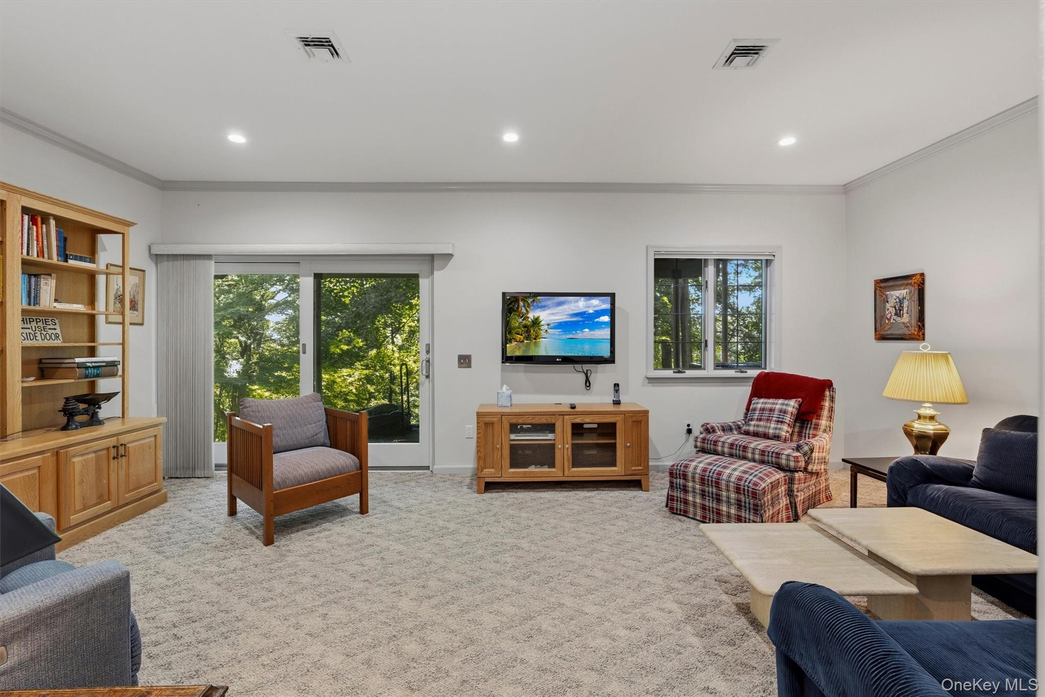 Furnished living room with built-in shelves, plaid chair, and sliding glass doors overlooking trees.