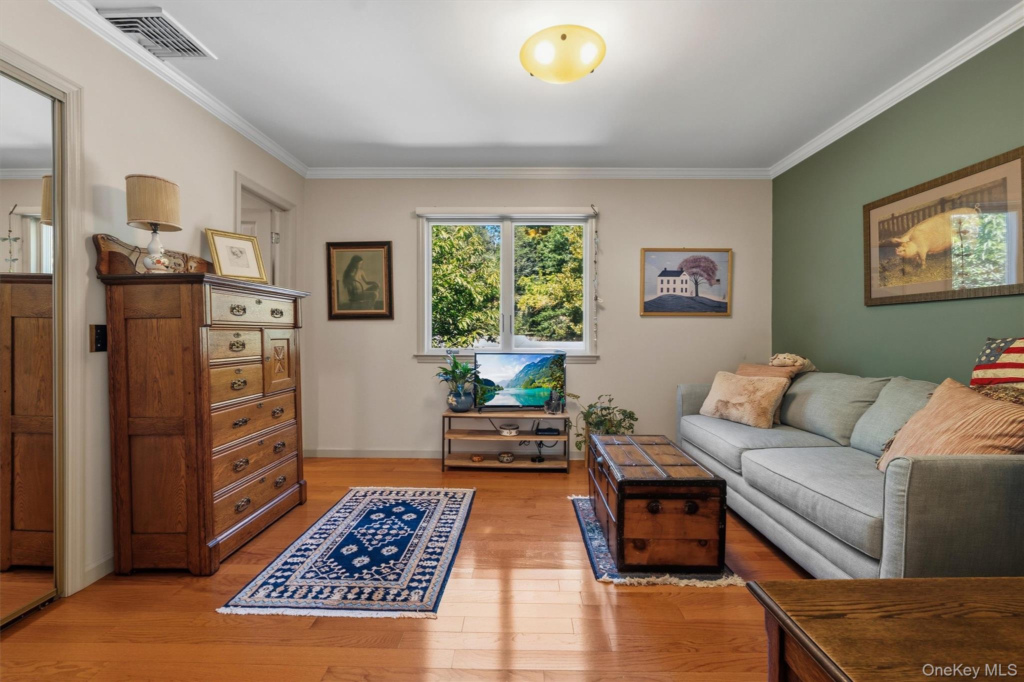 Living room with a gray sectional, antique dresser, patterned rugs, and a window overlooking leafy greenery.