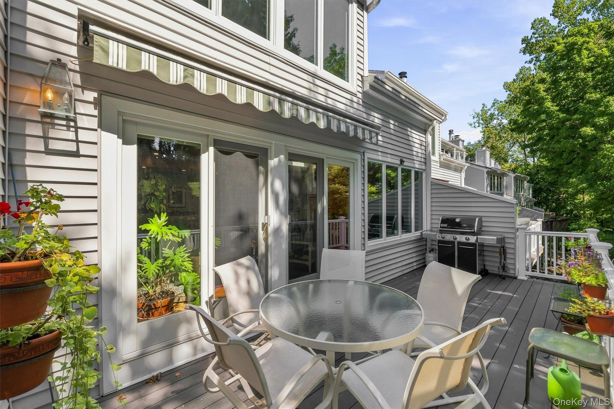 Furnished deck with a round glass-top table, potted plants, and gas grill beside a gray-sided home.
