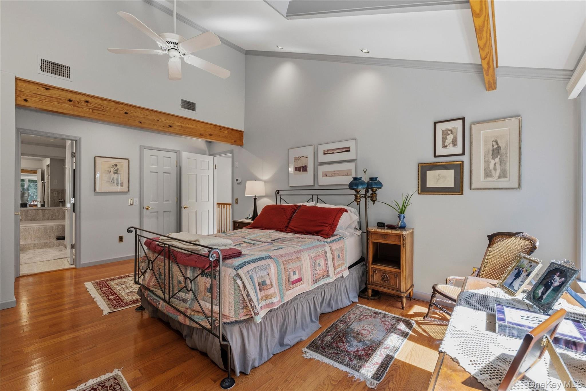 Furnished bedroom with a patchwork-quilted iron bed, vaulted ceiling, exposed beam, and hardwood floors.