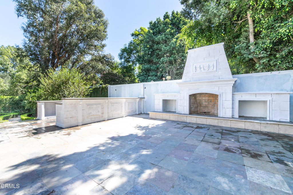 Stone patio with a white masonry fireplace and built-in openings, surrounded by mature trees.