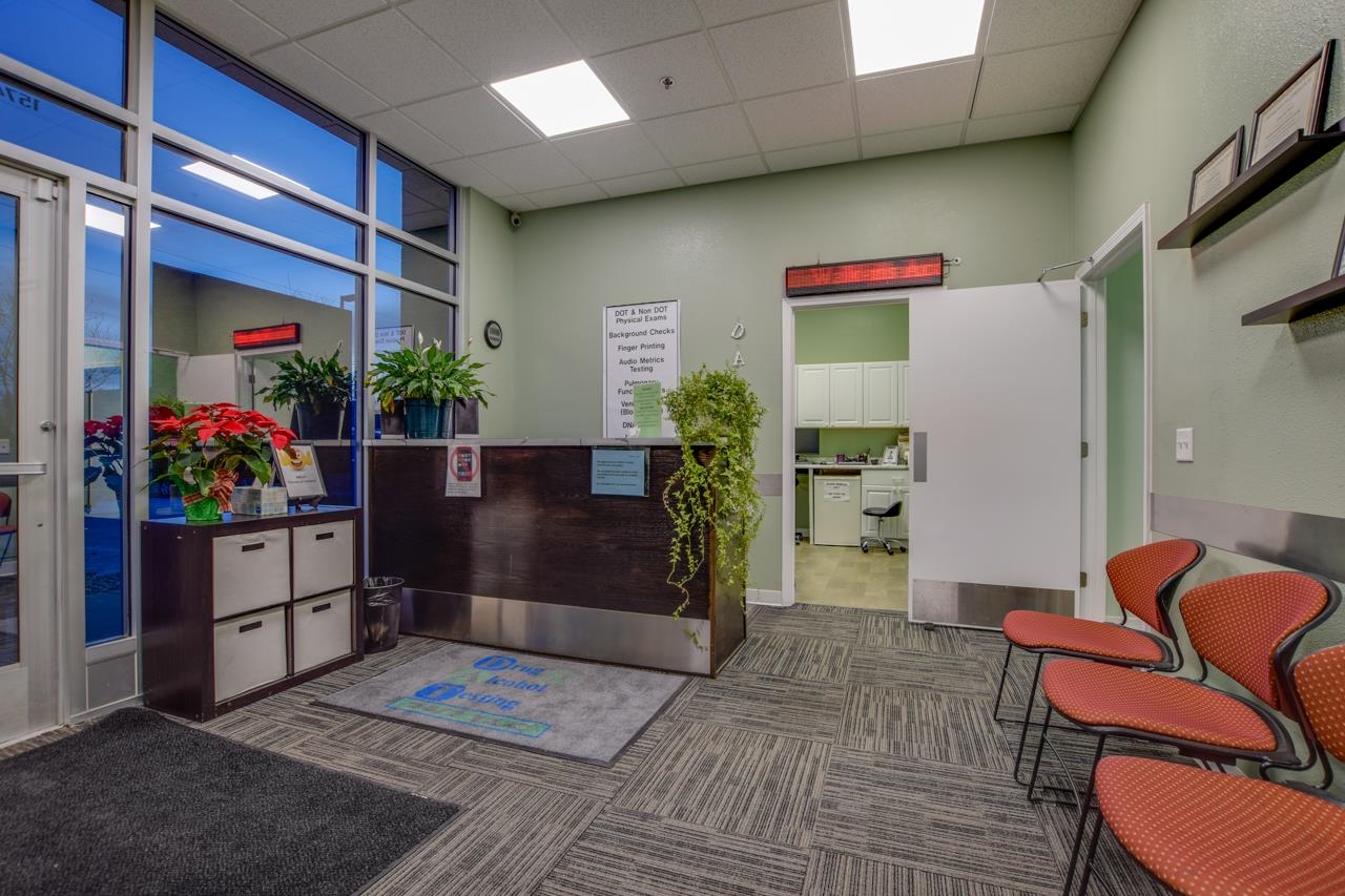 Office reception area with a front desk, red waiting chairs, large windows, and an open doorway to a break room.