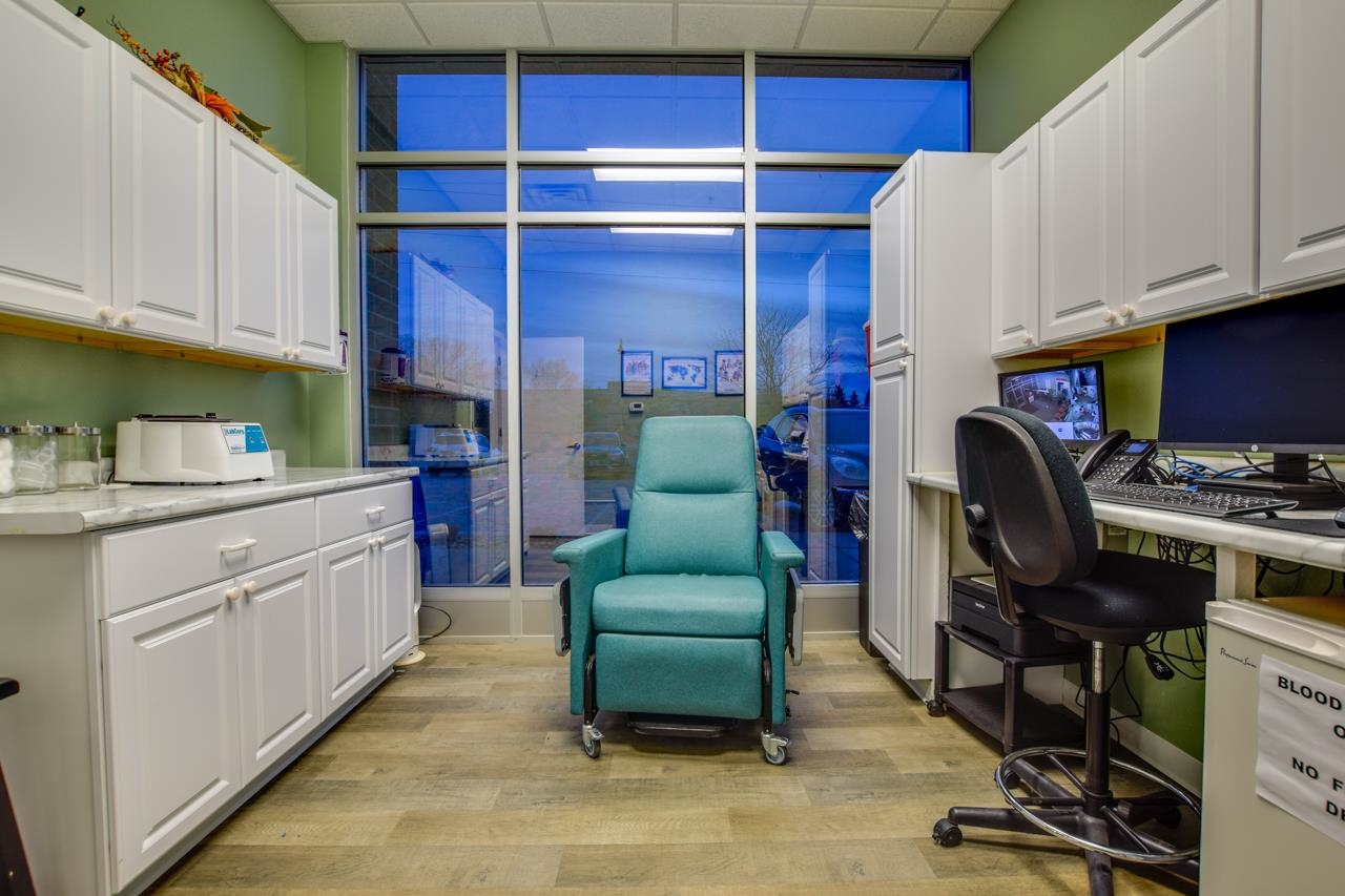 Medical treatment room with a teal reclining chair, white cabinetry, and a workstation beside large windows.