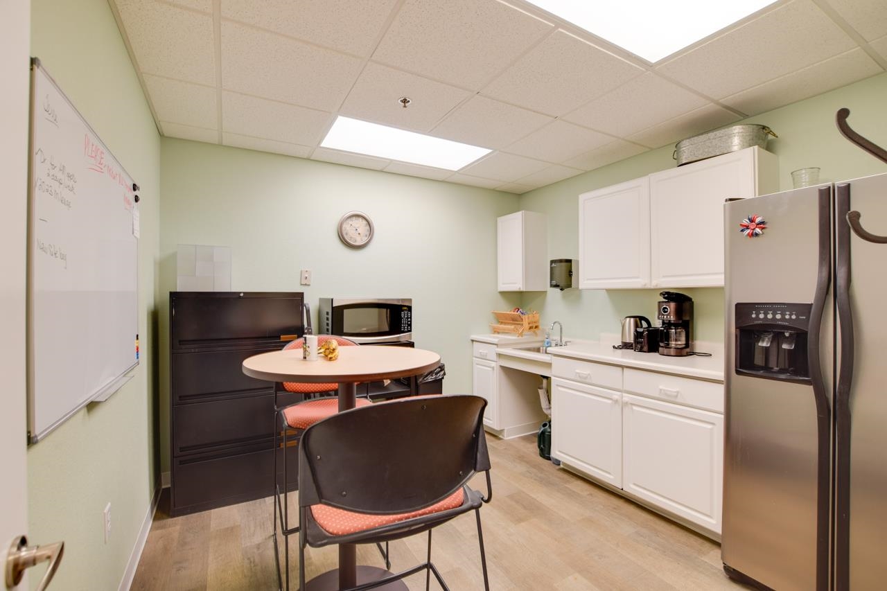 Kitchenette with white cabinets, stainless steel refrigerator, microwave, and a round table beside a whiteboard.