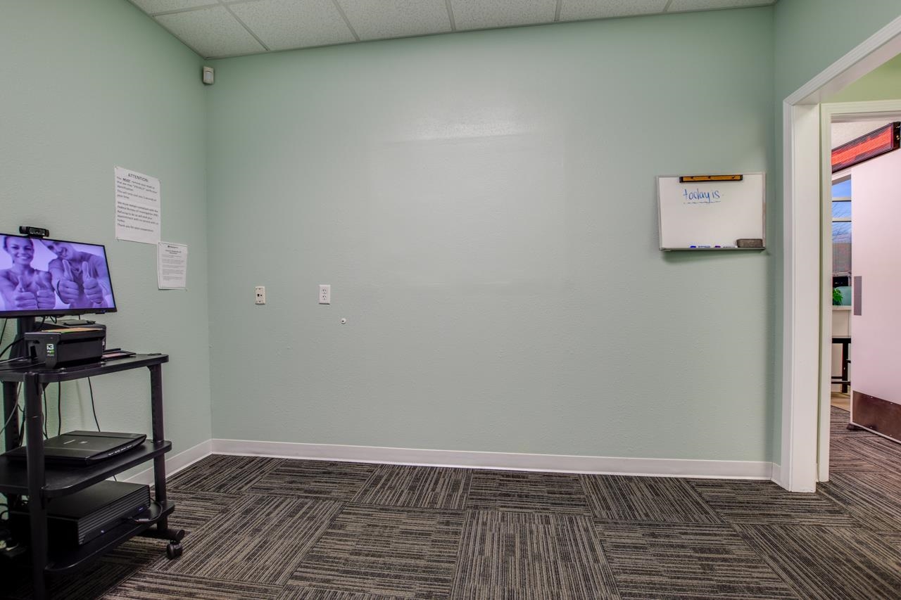 Office meeting room with pale green walls, patterned carpet, a whiteboard, and computer equipment.