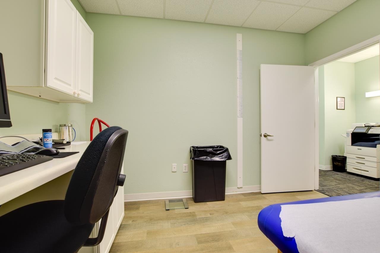 Treatment room with an exam table, desk, wall cabinets, and an open doorway to an adjoining office.