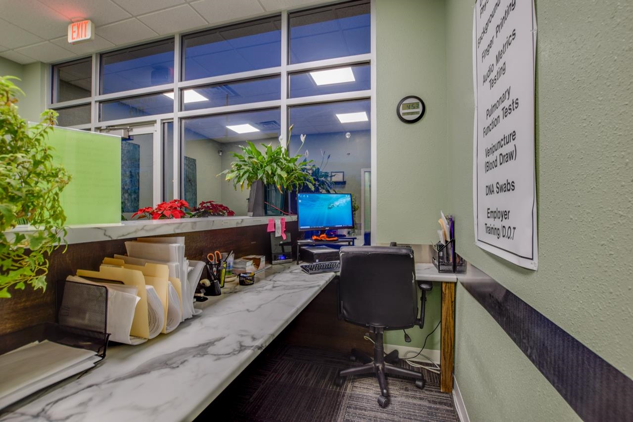 Office reception counter with a computer, rolling chair, glass partition, and medical testing services sign.