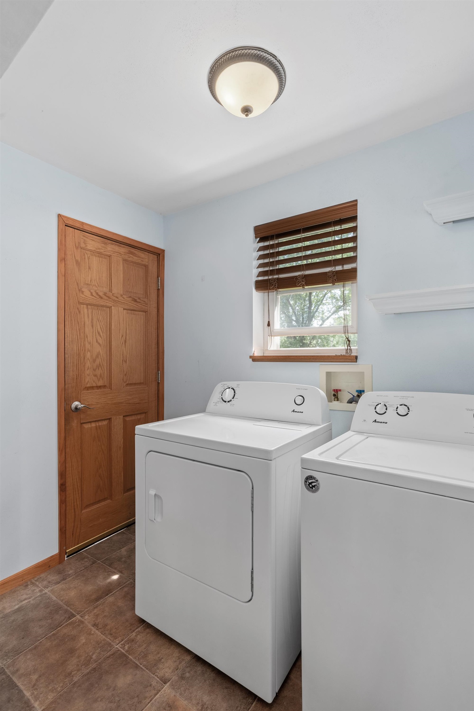 Laundry room with white washer and dryer, brown tile floor, and a window with wood blinds.
