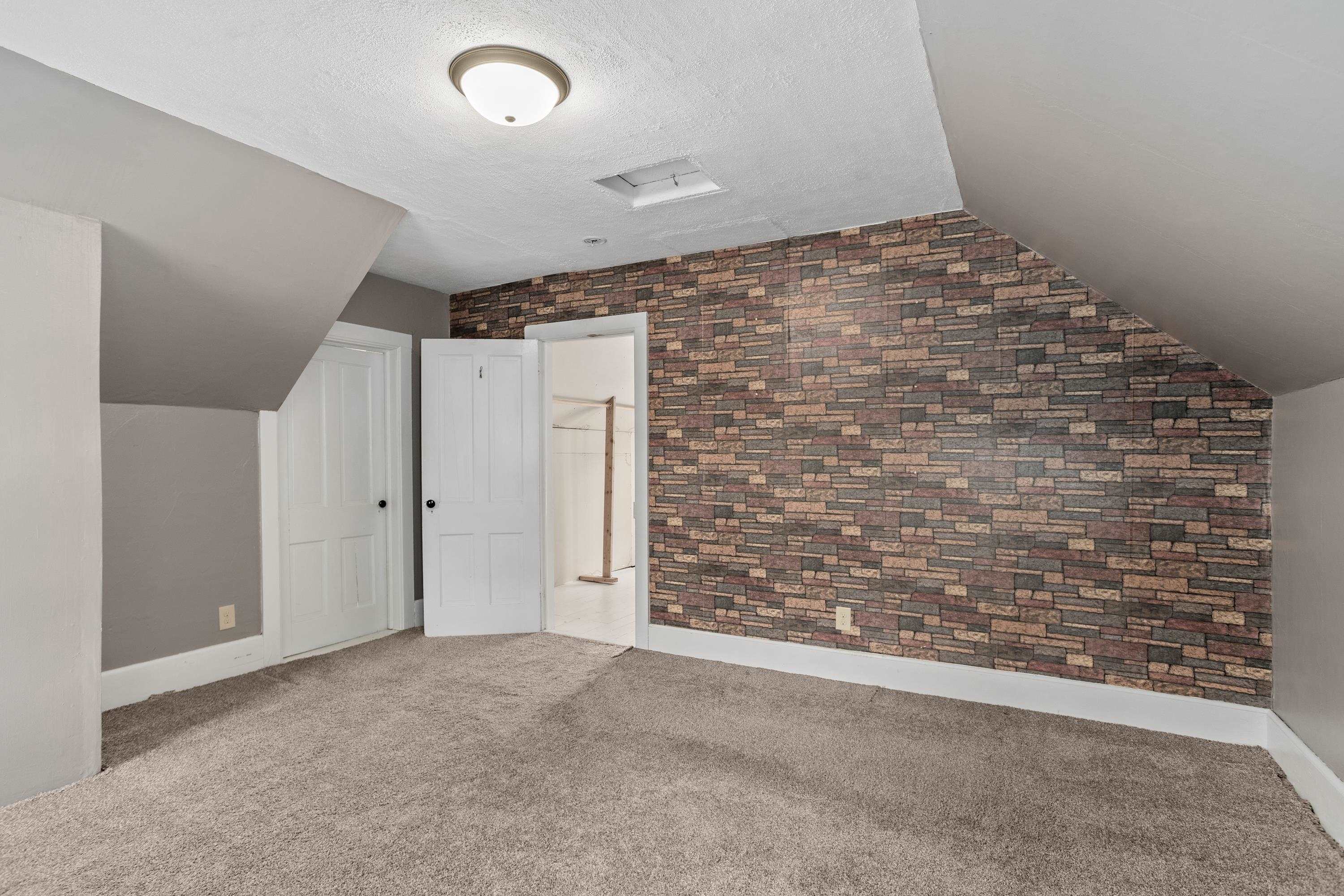 Empty carpeted room with a sloped ceiling and stone-pattern accent wall.