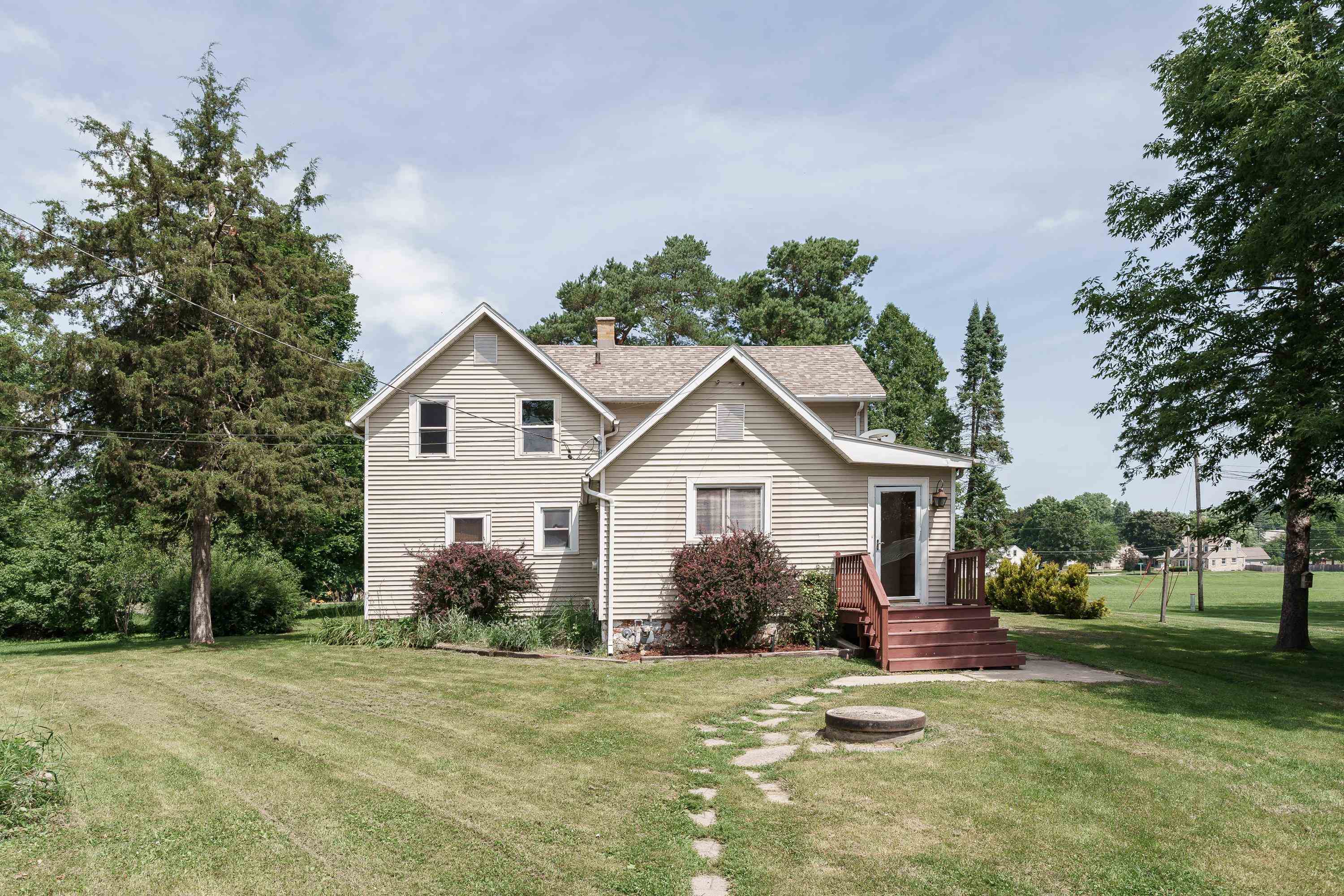 Two-story beige house with a small raised deck and steps, surrounded by a lawn, shrubs, and mature trees.