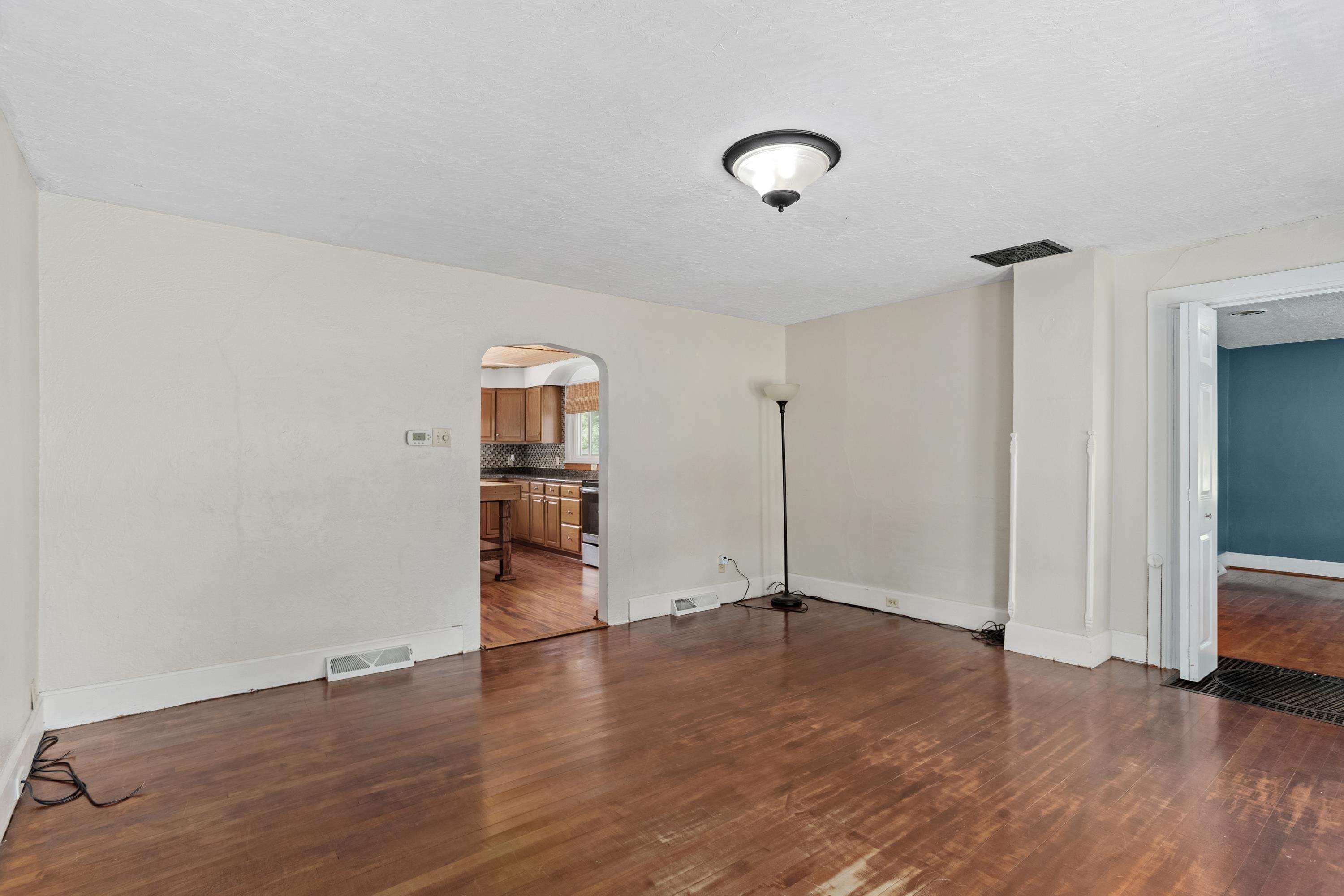 Empty living room with worn hardwood floors and an arched opening to the kitchen.