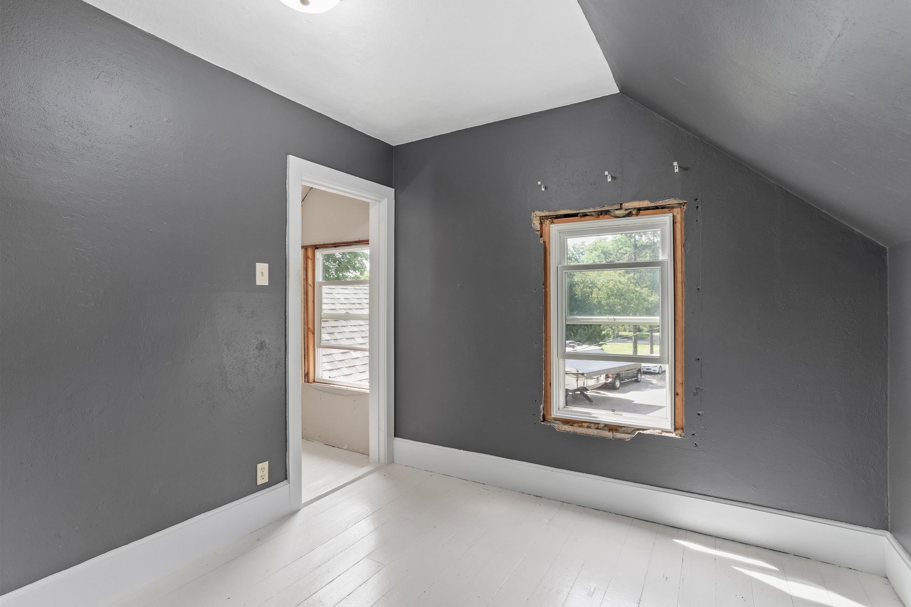 Empty attic room with gray walls, white floors, and exposed framing around a window.