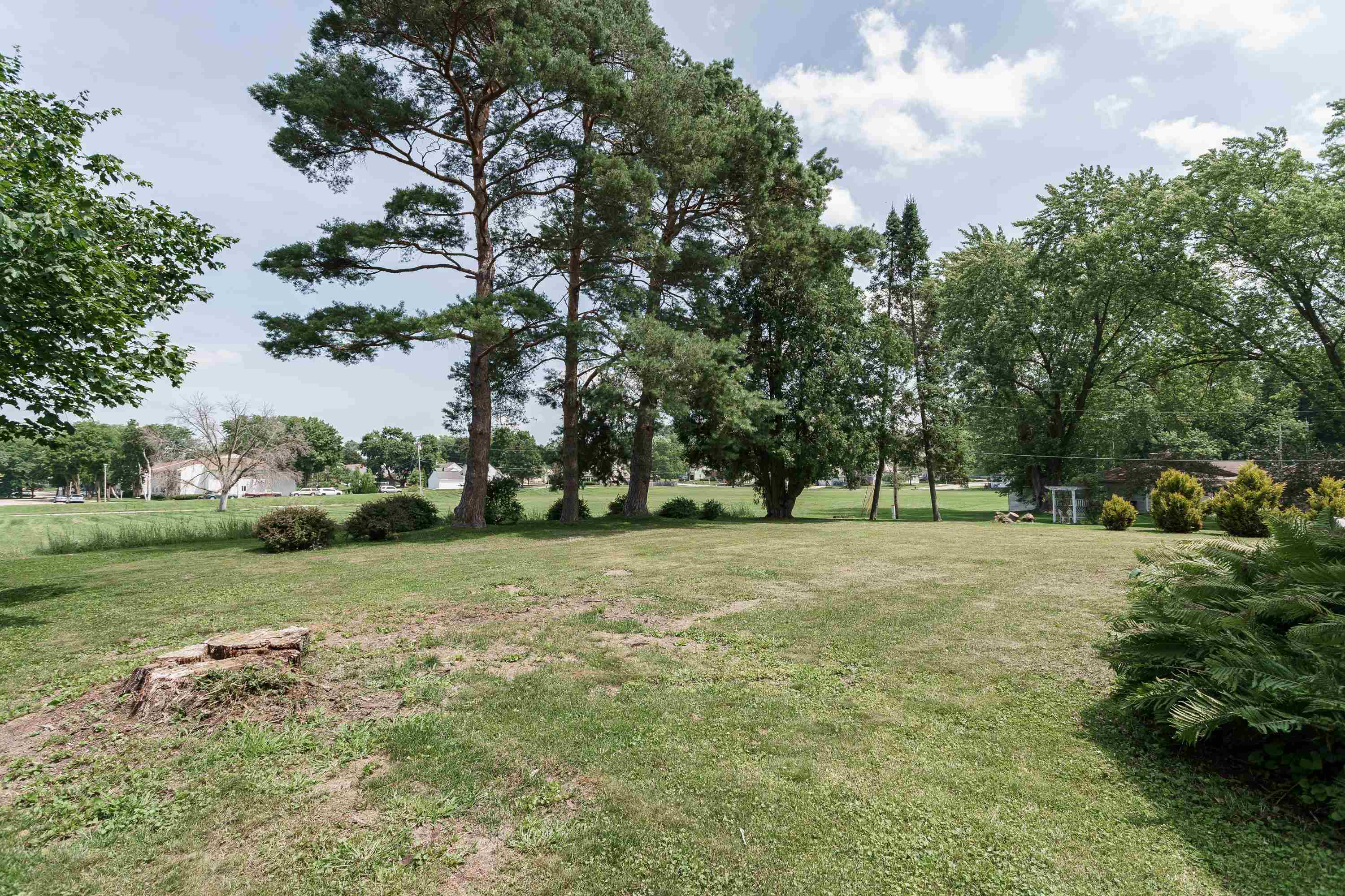 Spacious grassy yard with mature trees and a cut stump in the foreground.