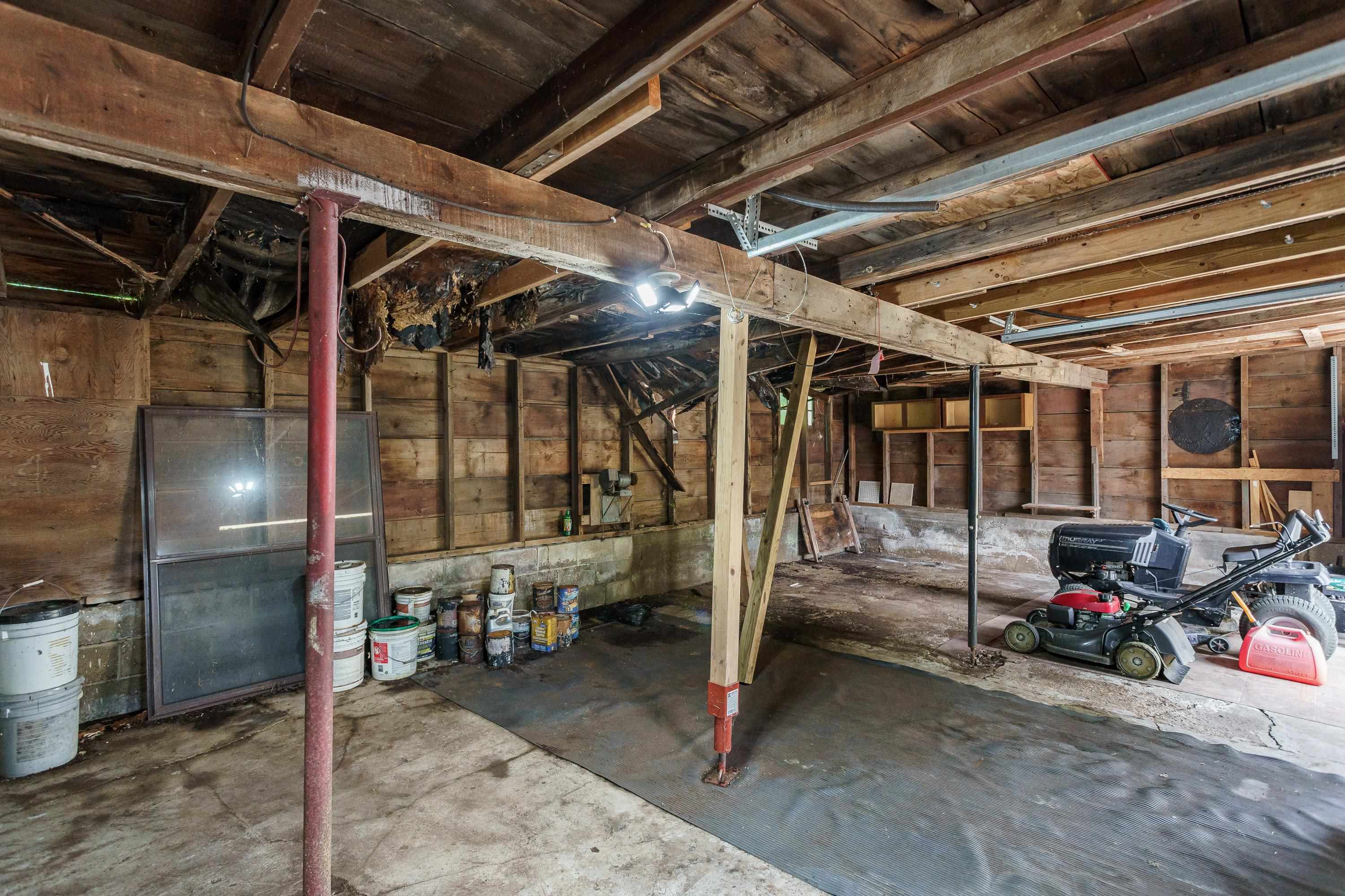 Unfinished garage with exposed beams, support posts, paint buckets, and a lawn mower.