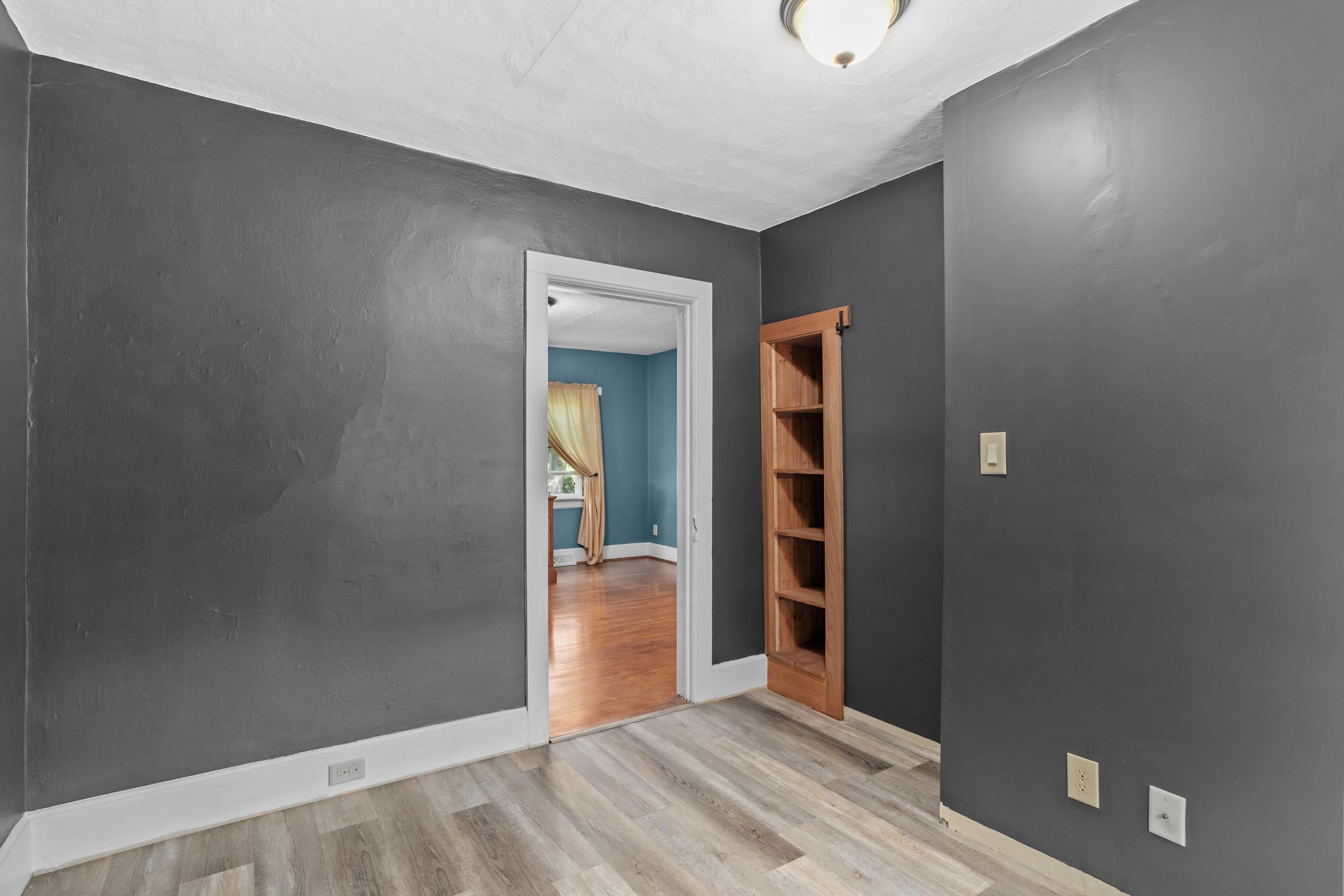 Empty gray room with wood-look flooring and built-in shelves beside a doorway to a blue room.