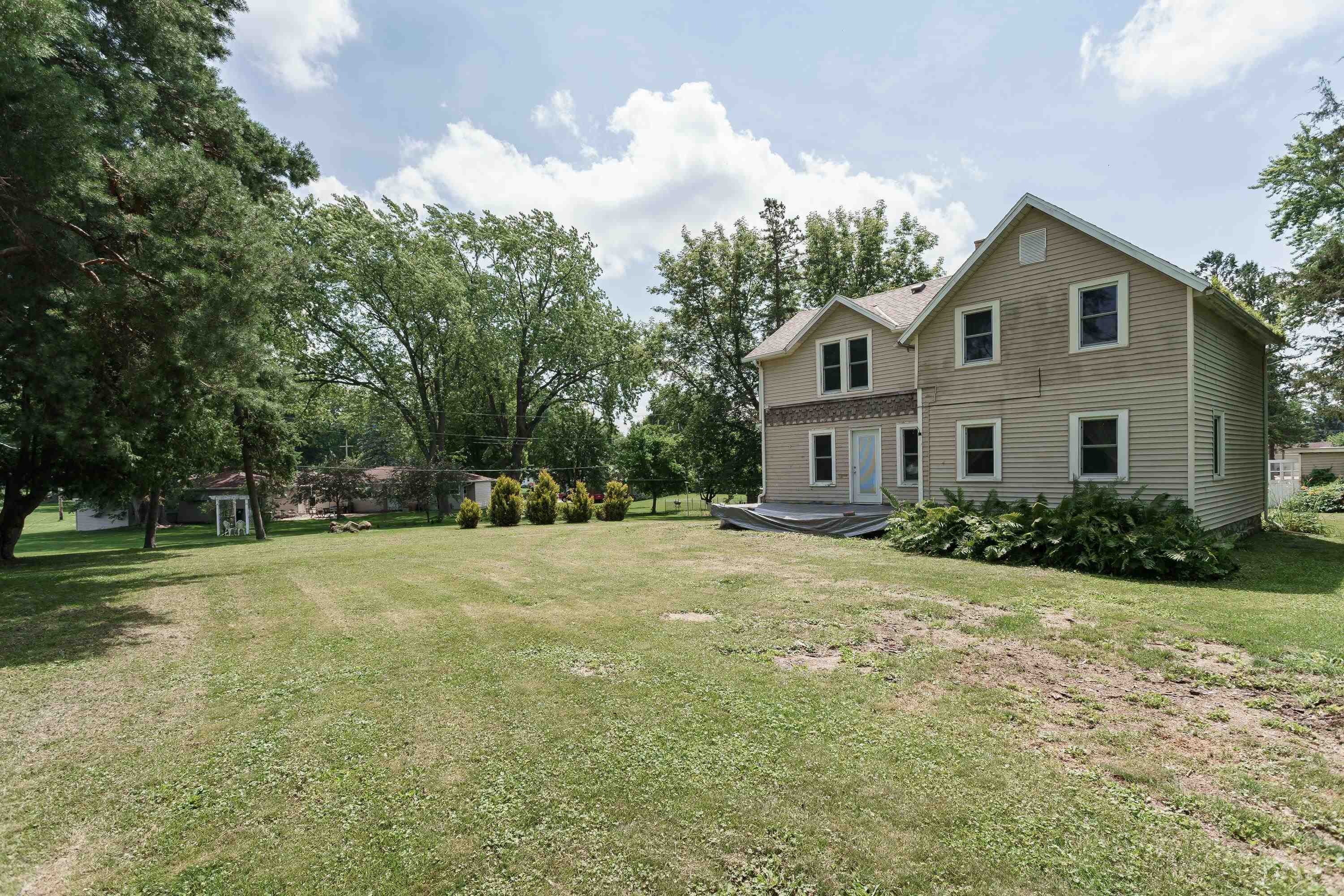 Two-story beige house beyond a broad lawn, framed by mature trees and shrubs.