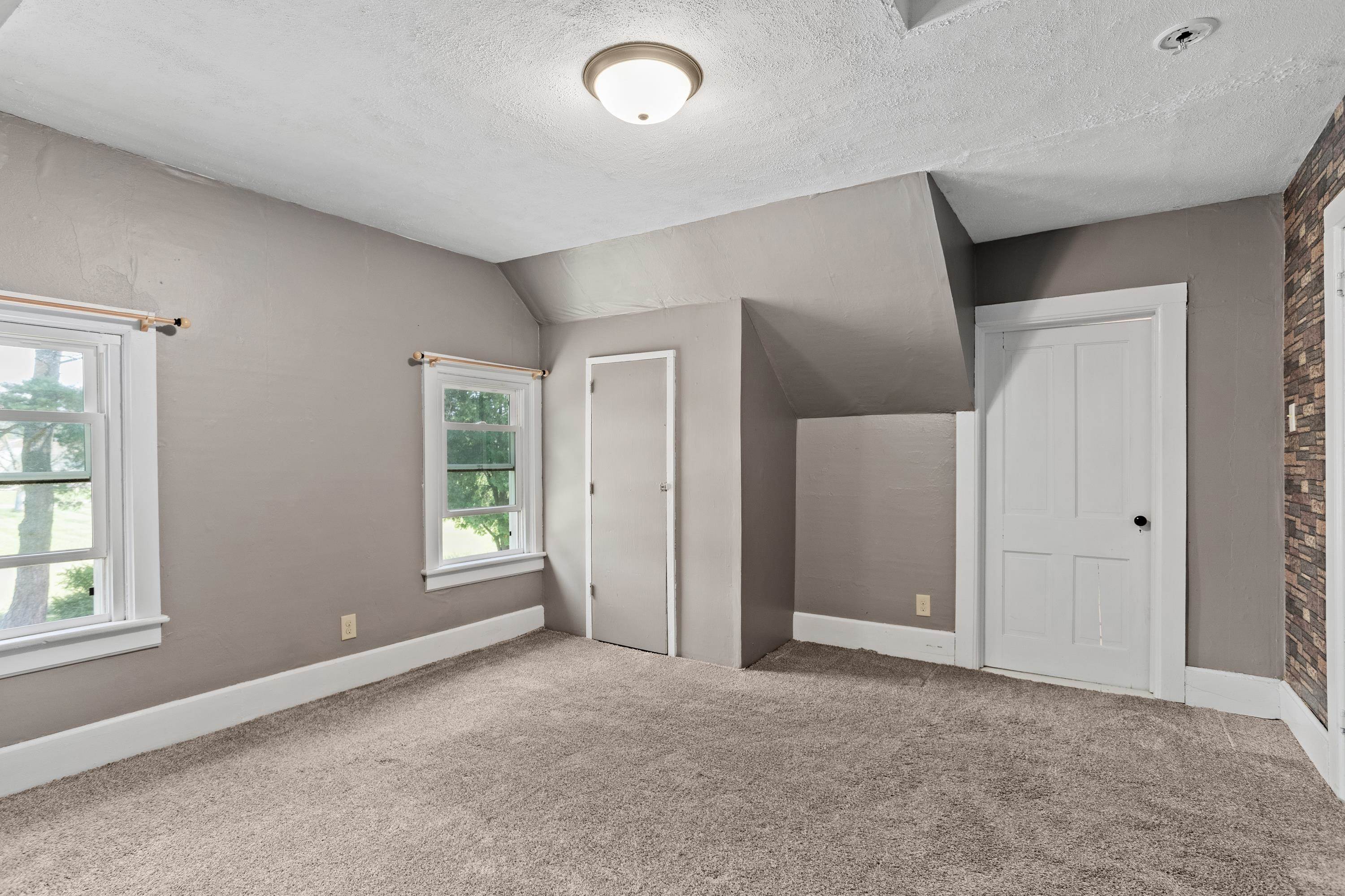 Empty carpeted room with gray walls, two windows, white doors, and a sloped ceiling.